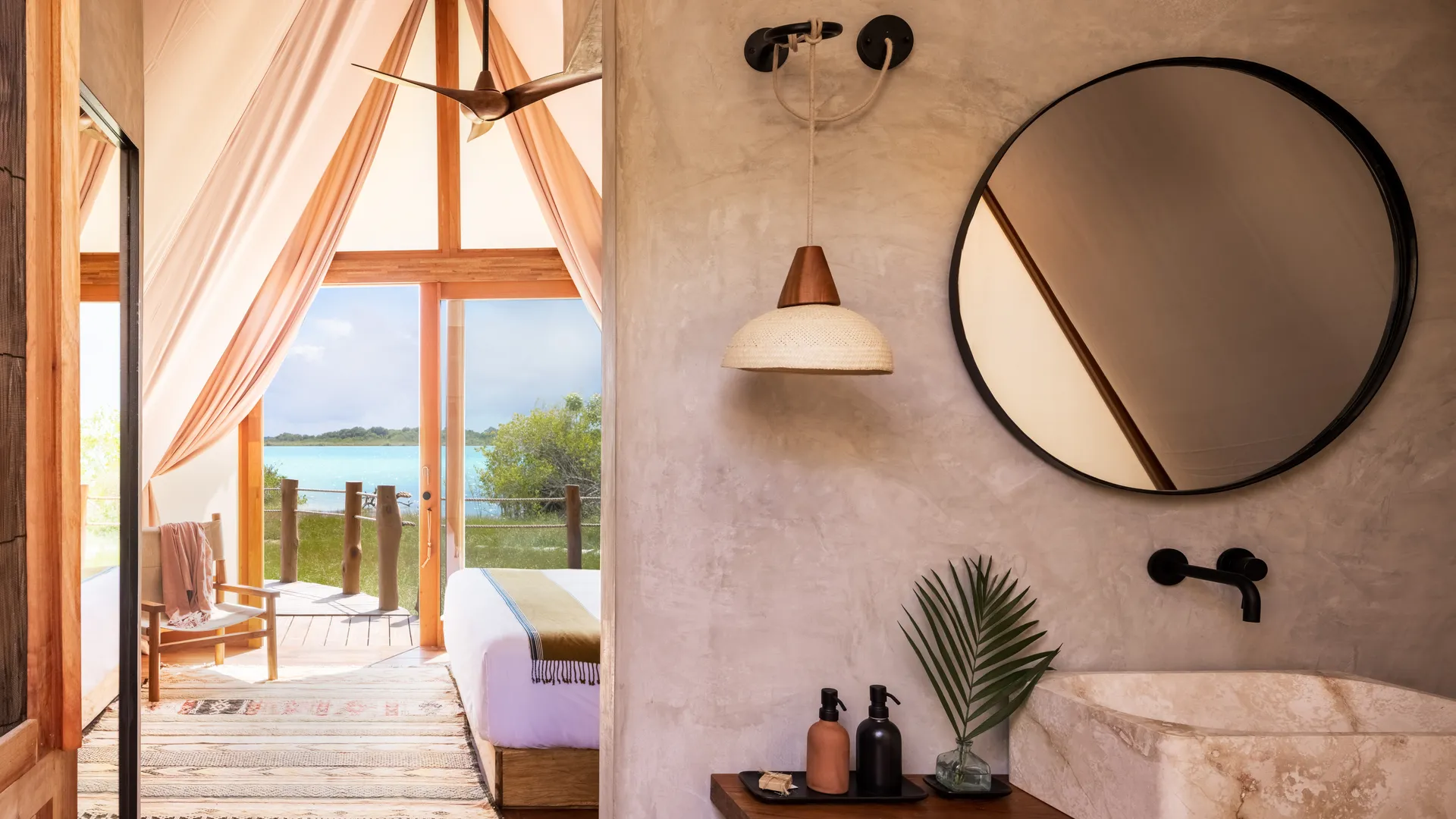 A stone-and-plaster bathroom with a round mirror beside a lagoon-view bedroom at Our Habitas Bacalar