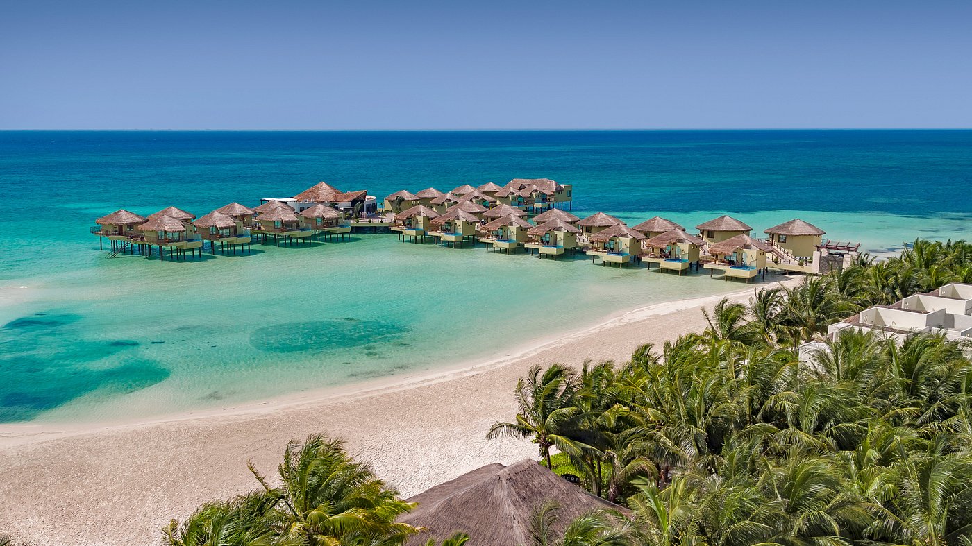 Aerial of thatched overwater bungalows on stilts above turquoise water at Palafitos, Maroma Beach